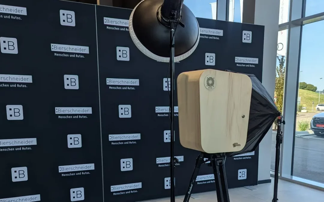 Fotobox mit Holzgehäuse, Stativ, Softbox und Ringlicht vor schwarzer Werbewand mit Logo und Schriftzug „Bierschneider – Menschen und Autos“ im Firmenevent / Mitarbeiterfeier / Sommerfest mit großen Glasfenstern.