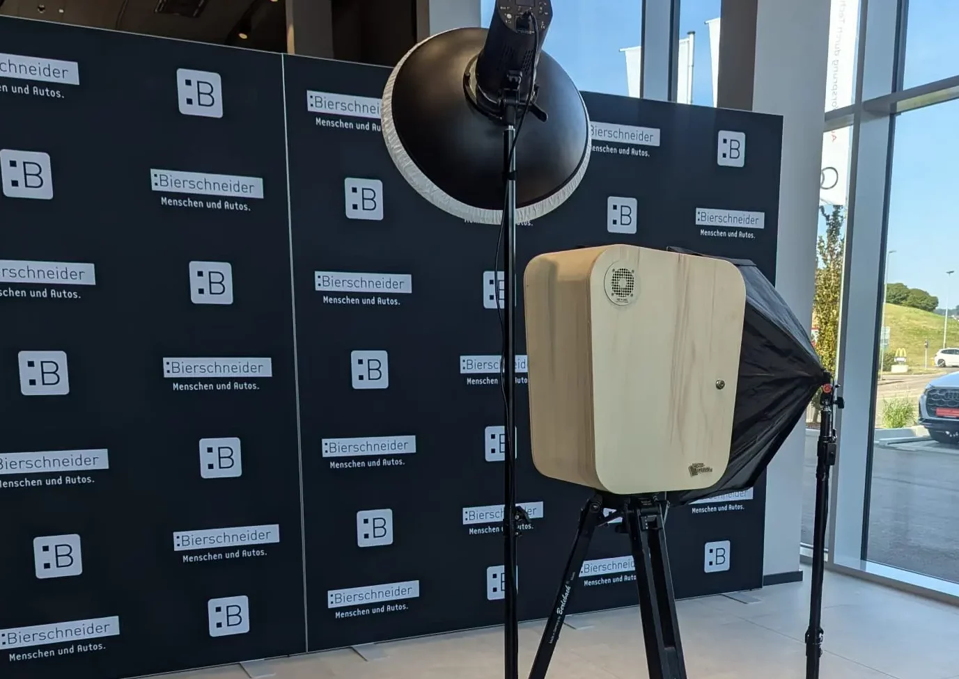 Fotobox mit Holzgehäuse, Stativ, Softbox und Ringlicht vor schwarzer Werbewand mit Logo und Schriftzug „Bierschneider – Menschen und Autos“ im Firmenevent / Mitarbeiterfeier / Sommerfest mit großen Glasfenstern.